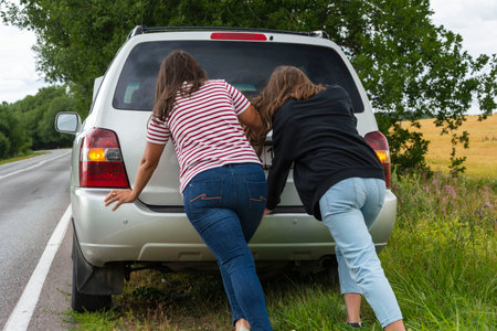 A broken car. Two young women push a broken car on the road, a breakdown, against the background of a yellow field.の写真素材