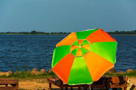 A beach umbrella from the sun and a wooden chaise longue on the beach, sunlight, water and sand. The beach of the tourist base on the lake.の写真素材