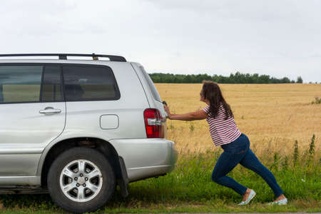 A broken car. A young woman pushes a broken car on the road, a breakdown, against the background of a yellow field.の写真素材