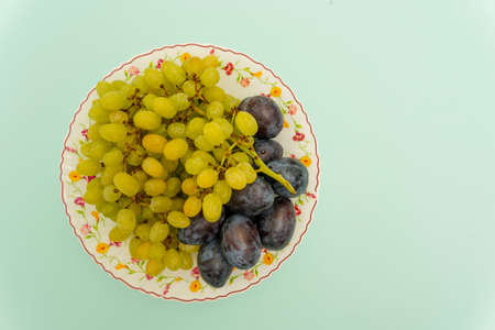 Ripe organic white grapes with round blue organic plums on a plate on a turquoise background, fruit, healthy food, vegetarianism. Top view. Vegan foodの写真素材