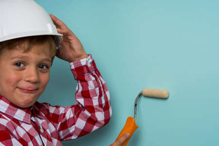 A cute boy in a plaid shirt and a construction helmet with a paint roller in his hands paints a wall, a portrait of a small builder in a helmet.の写真素材