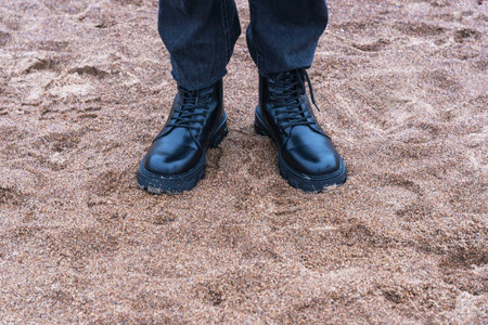 Feet in black leather rough boots and black fashionable denim pants standing on the sand on the...
