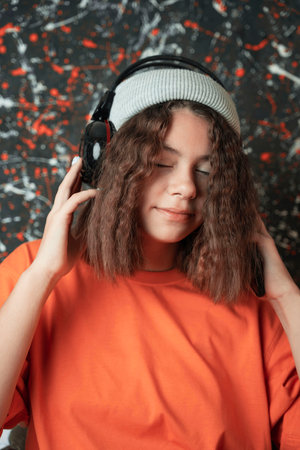 A cute European teenage girl in a fashionable hat with beautiful wavy hair listens to music through large headphonesの写真素材