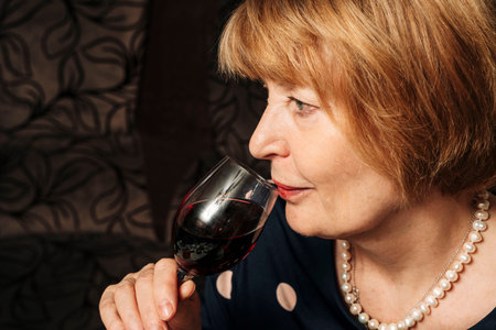 A pretty woman aged drinks wine from a glass sitting in the living room on a home sofaの写真素材