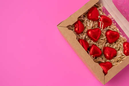 A group of heart-shaped chocolates wrapped in red foil, lying in a gift box on a pink backgroundの写真素材