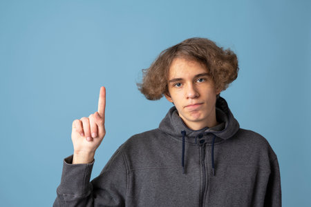 a teenager in a gray hoodie with wavy hair points up with his index finger on a blue background