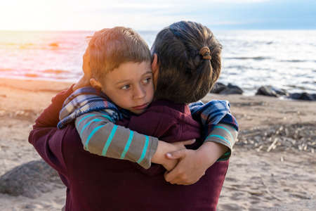 Cute sad preschool boy hugs his dad on the seashore in sunset lightの写真素材