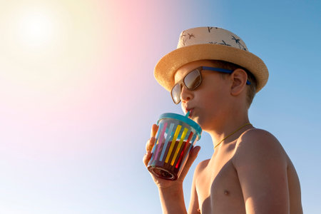 happy boy in hat and sunglasses drinking juice on the beachの写真素材