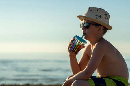 happy boy in hat and sunglasses drinking juice on the beachの写真素材
