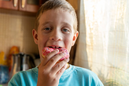 a boy eats a pink donut in the home kitchen. a child is having fun with a donut