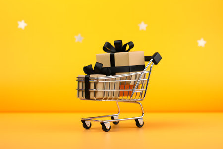 a supermarket cart with a gift box tied with a ribbon. Yellow background. a shopping basket full of gift boxes with ribbons on a yellow background. Space for copying. The concept of sales, discounts, purchases and deliveryの素材