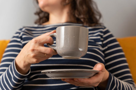 woman in a striped sweater sits on the sofa and holds a cup of coffee in handsの写真素材