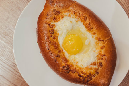 A close-up of a freshly baked khachapuri, a classic Georgian egg and cheese bread, on a rustic table in a charming cafe. The golden crust and gooey cheese are incredibly tempting.の写真素材