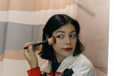 A teenage girl is sitting in front of a mirror, applying makeup with a soft brush. She is focused while enhancing her features using the beauty product.の写真素材