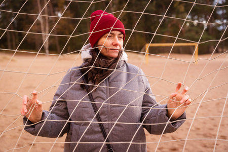 A stylish young woman is posing by a football goal net on a sandy beach, wearing a fashionable jacket and hat, showcasing the latest autumn fashion trends in a picturesque setting.の写真素材