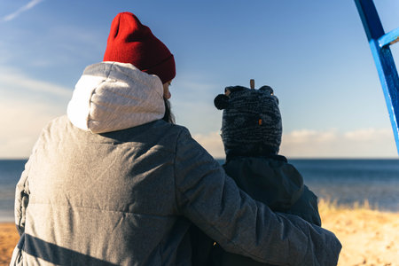 A woman in an autumn jacket and her child are sitting on a bench on a deserted autumn beach on a sunny day. Family relations. Motherhood. Active rest on a day off. healthy lifestyleの写真素材