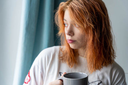 A cheerful red-haired teenage girl from Generation Z is standing by a window, holding a hot beverage and delighting in the cozy warmth as she gazes outside.の写真素材