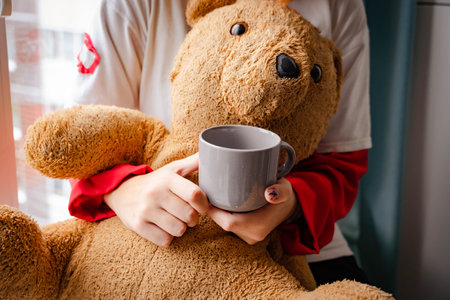 Close-up shot capturing the hands of a teenage girl from generation Z as she enjoys a warm cup of tea or coffee while sitting beside a window, embracing a teddy bear for comfort.の写真素材