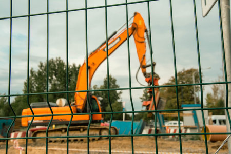 An industrial construction site with an orange excavator in operation, viewed through the metal grating of the surrounding fence. The heavy machinery and activity convey the work in progress.の写真素材