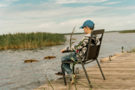 On the quiet lakeshore, a school-age boy finds solace as he sits alone on a chair, contemplating the stillness of nature's serene beauty, immersing himself in tranquil surroundings.の写真素材
