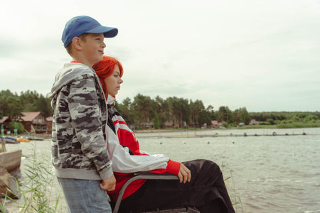 redheaded Generation Z girl and her younger brother relaxing by the lake, showcasing the strong bond and affection within the family in a beautiful outdoor setting.の写真素材