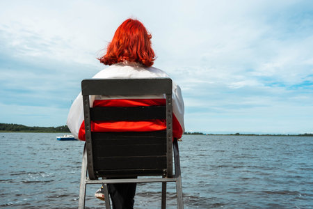 A contemplative Generation Z redhead girl sitting by a serene lake, surrounded by nature's beauty, enjoying a peaceful moment of solitude and reflection.の写真素材