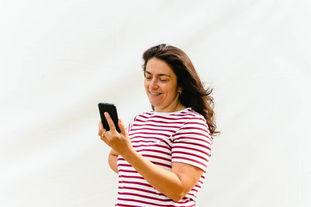 Stylish Middle-Aged Woman in Striped Blouse Using Smartphone on Bright Backgroundの写真素材