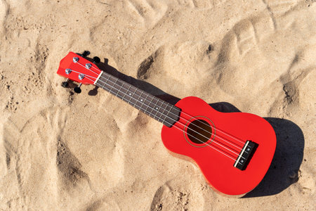 A red ukulele laying in the sandy beach under the sun