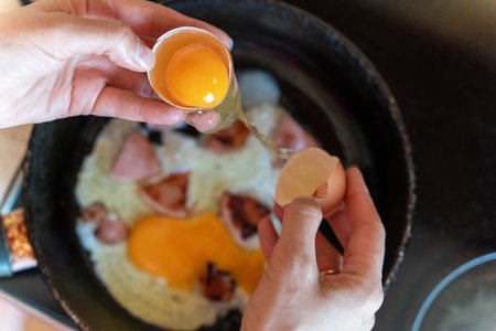 A person cracks an egg into a pan of scrambled eggs and ham