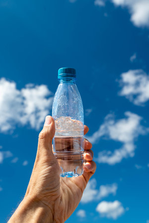 Hand lifts a clear water bottle towards a sunny sky with cloudsの写真素材