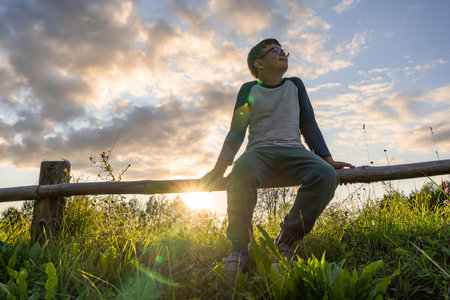 Boy sitting on a fence, watching the sunset over grassy fieldsの写真素材