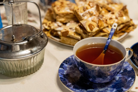 Porcelain teacup filled with tea sits on a saucer with a spoon, beside a metal kettle and a plate of cake, creating a cozy teatime sceneの写真素材