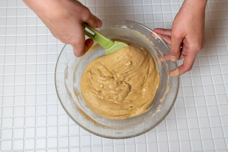 Hands mixing cake batter in a glass bowl with a green spatula, over white tiled kitchen counterの写真素材
