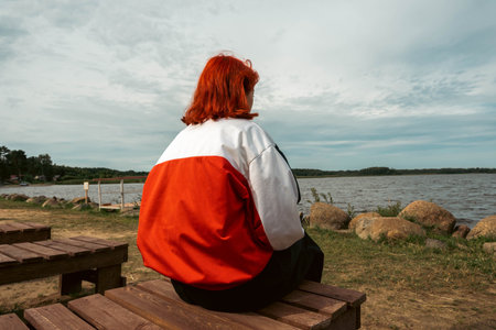 A person with red hair sits on the shore of a tranquil lake, gazing out at the serene water with...