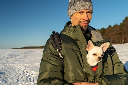 Man wearing winter clothes holding small white dog in jacket pocket in snowy landscapeの写真素材