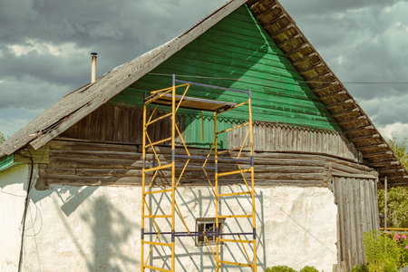 Scaffolding standing next to old wooden house under cloudy sky during renovationの写真素材