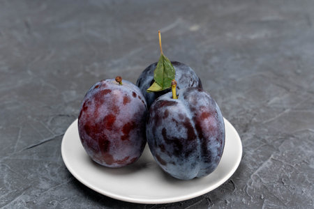 Three ripe blue plums in white saucer on grey background, fresh seasonal fruitsの写真素材