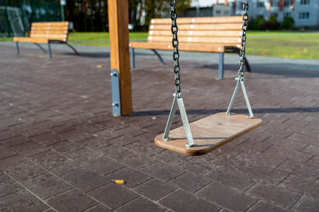 Wooden swing hanging from a sturdy frame in a playground, surrounded by benches and green grass,...