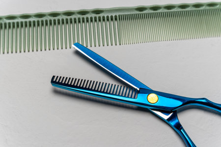 Professional hairdressing scissors with a blue handle positioned beside a green comb on a smooth...