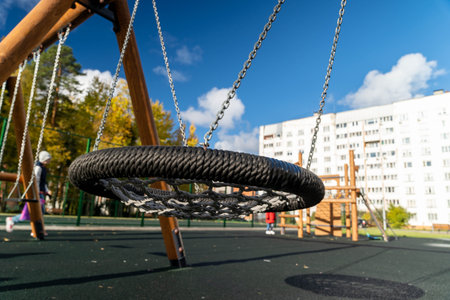 Circular swing made of durable material hangs from wooden frame in a vibrant playground, surrounded by trees and a modern building, inviting children to play and exploreの写真素材