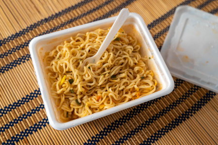 Instant noodles in a white takeout container with a plastic fork, placed on a bamboo mat, showcasing a delicious meal ready to enjoy with vibrant colors and texturesの写真素材