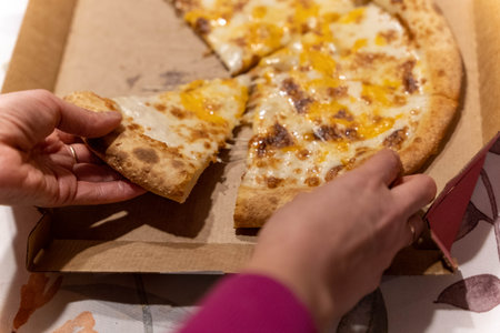 Hands reach for cheese pizza in a cardboard box on a table during dinner timeの写真素材