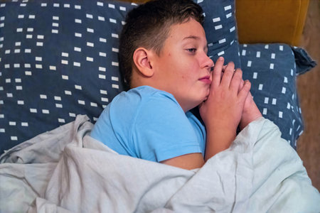 Child lying in bed with hands clasped together in a cozy room at nightの写真素材