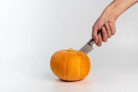 Hand holding a knife is positioned above a bright orange pumpkin on a white background, preparing for cutting or carving during autumn or Halloween festivitiesの写真素材