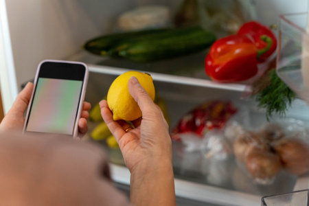 Person holding a lemon while using a smartphone in front of an open refrigerator filled with fresh...
