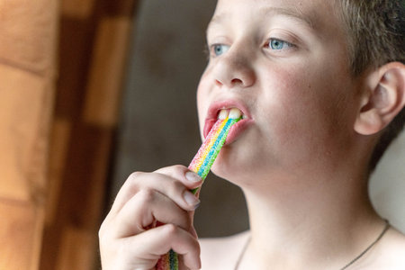 Young boy with light brown hair is enjoying a colorful candy stick while standing indoors near a...