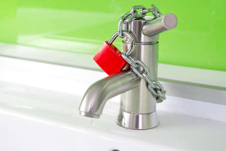 a waterdrop is dripping out of a modern faucet, locked by a red lock and chainの写真素材