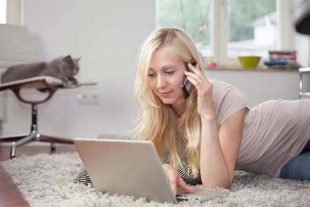 Relaxed woman is using the laptop and telephone in her livingroom, lying on the carpetの写真素材