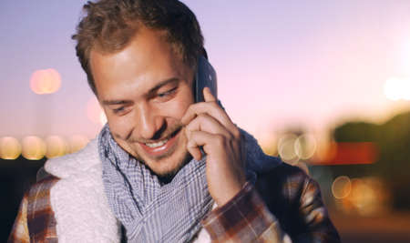 Handsome young man speaking on smart phone at autumn sunset in city. Using smartphone for a phone call, smiling happy wearing urban hipster outfit outdoors at dawn.の写真素材