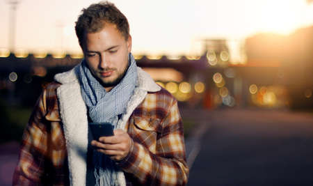 Handsome young man sms texting using app on smart phone at autum Summer sunset in city. Using smartphone smiling happy wearing urban hipster outfit outdoors.の写真素材